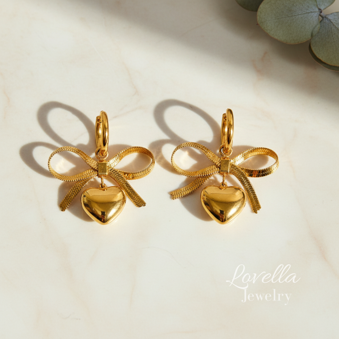 Gold bow-shaped earrings with heart pendants on a light background, featuring Lovella Jewelry branding.